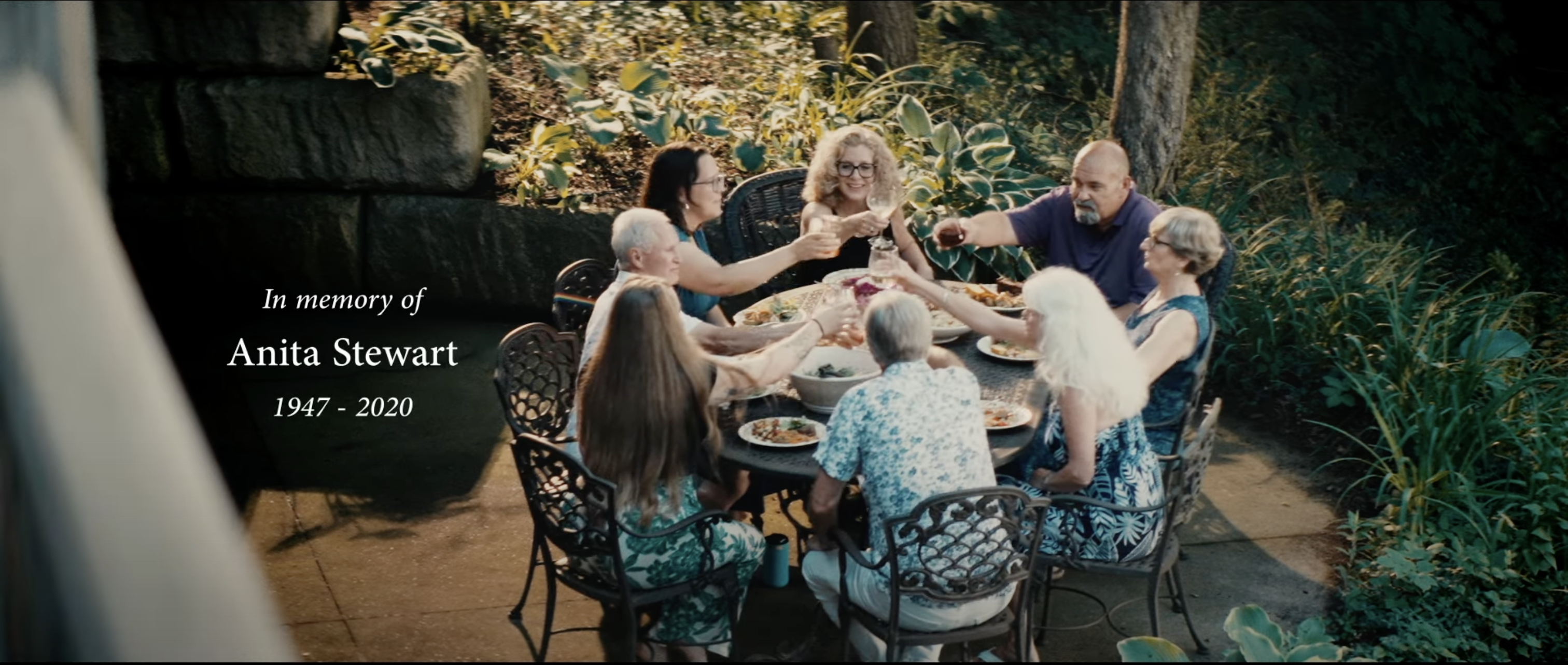 A group of seven people sitting outside at a table toasting their glasses. Text beside them reads "In memory of Anita Stewart (1947 - 2020)"